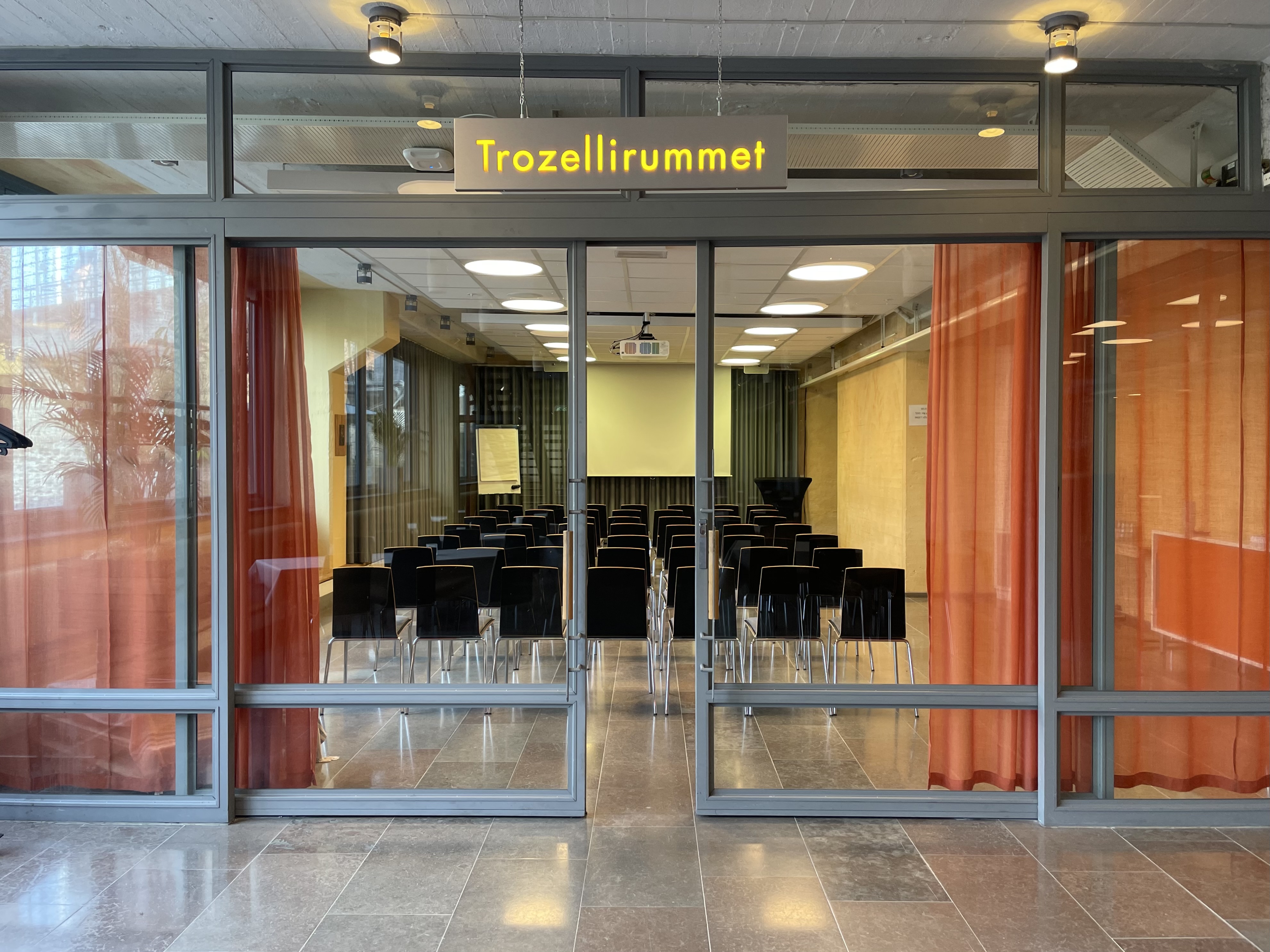 Konferensrum med stolar uppställda, fönstervägg och skylt med texten "Trozellirummet"