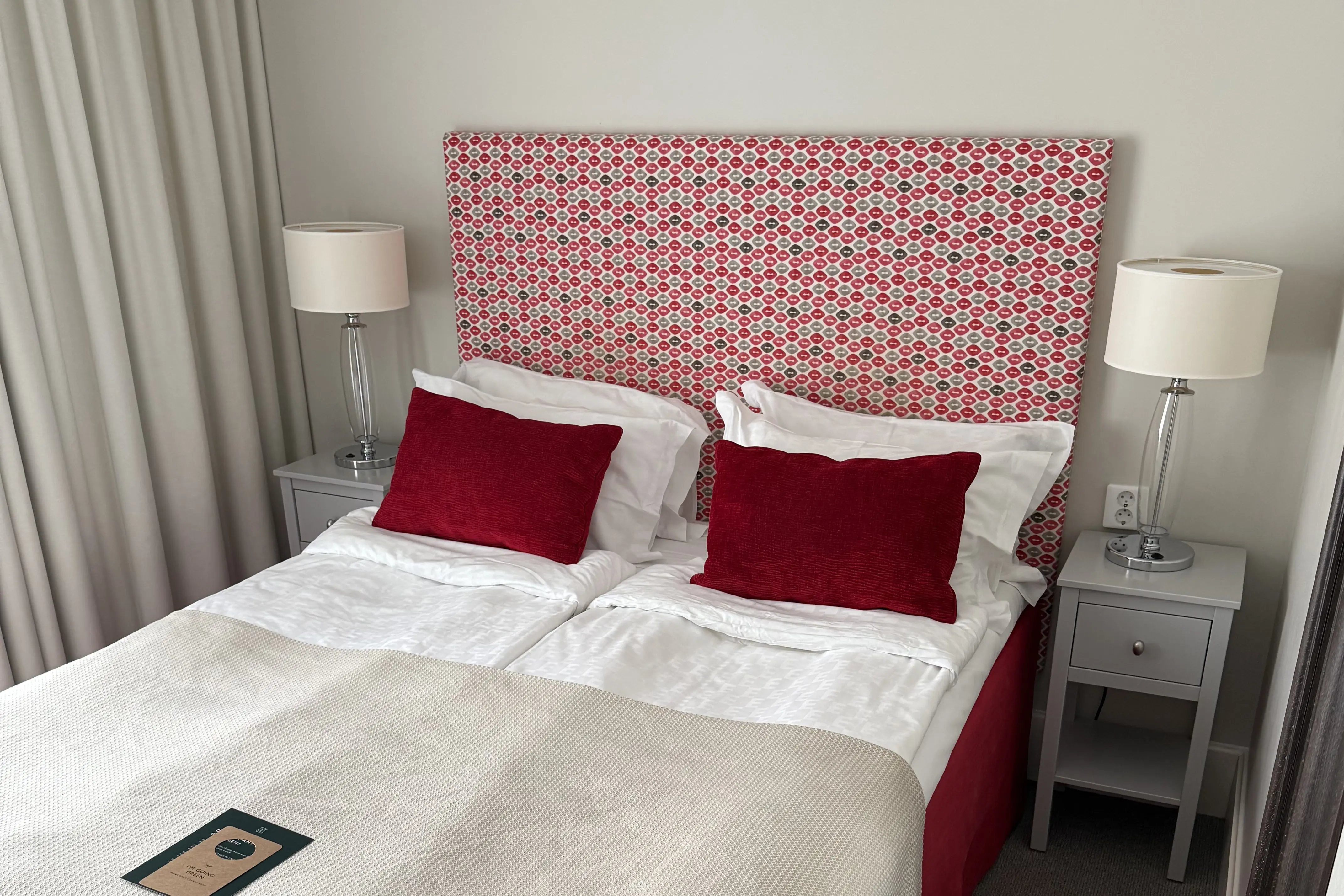 Cozy hotel room with twin beds featuring red patterned headboards, white bedding, and red accent pillows. Modern bedside tables with glass lamps add elegance.
