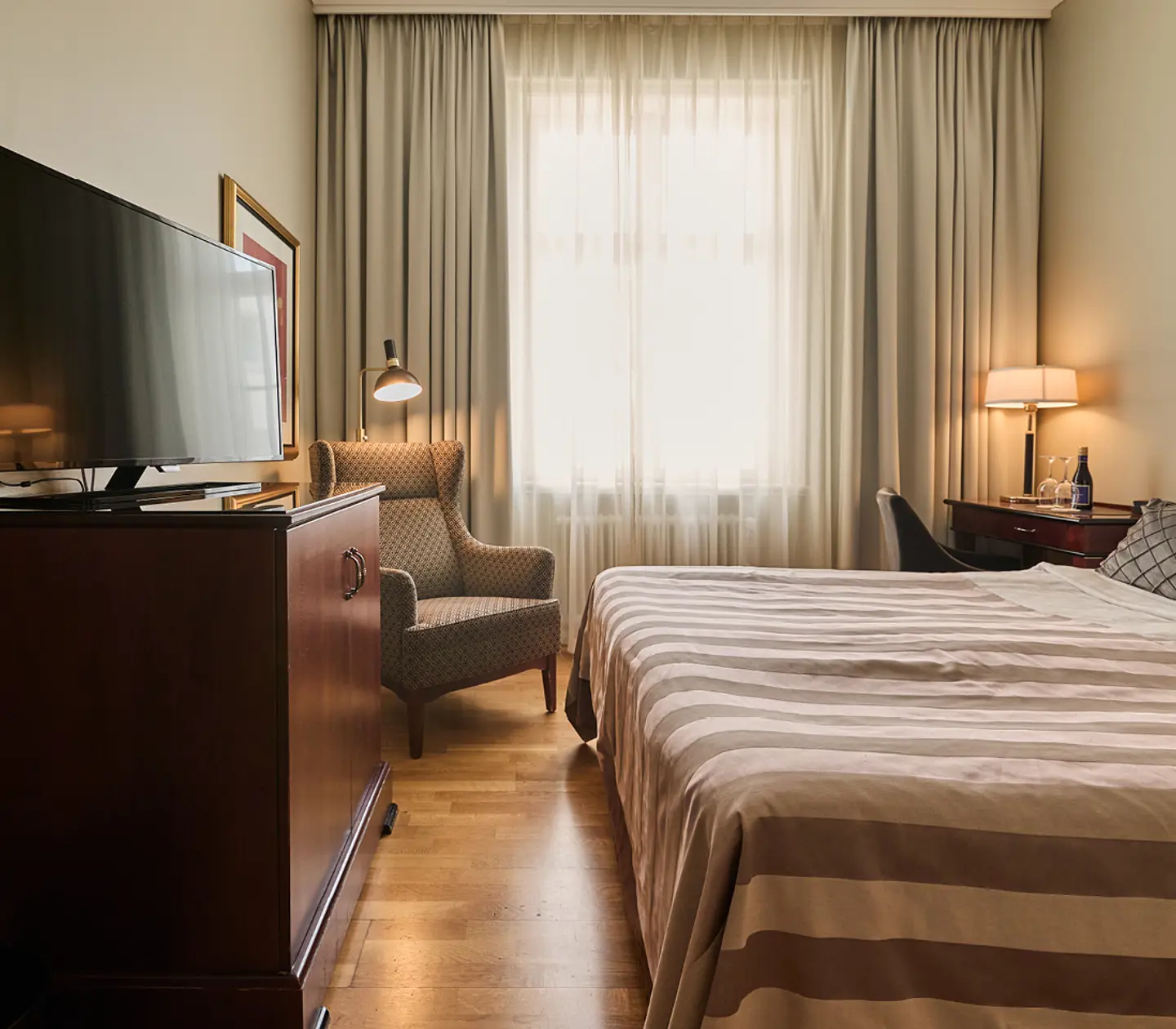 Cozy hotel room featuring a double bed with a decorative floral headboard, red accent pillows, and a beige blanket, accompanied by bedside tables with modern lamps.
