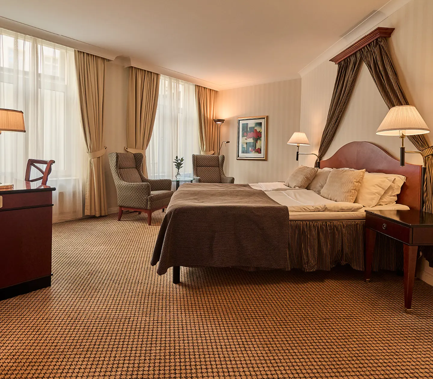 Modern hotel room with a cozy bed, stylish gray armchair, comfortable sofa, and round glass coffee table, accented by framed art and large windows with beige curtains.