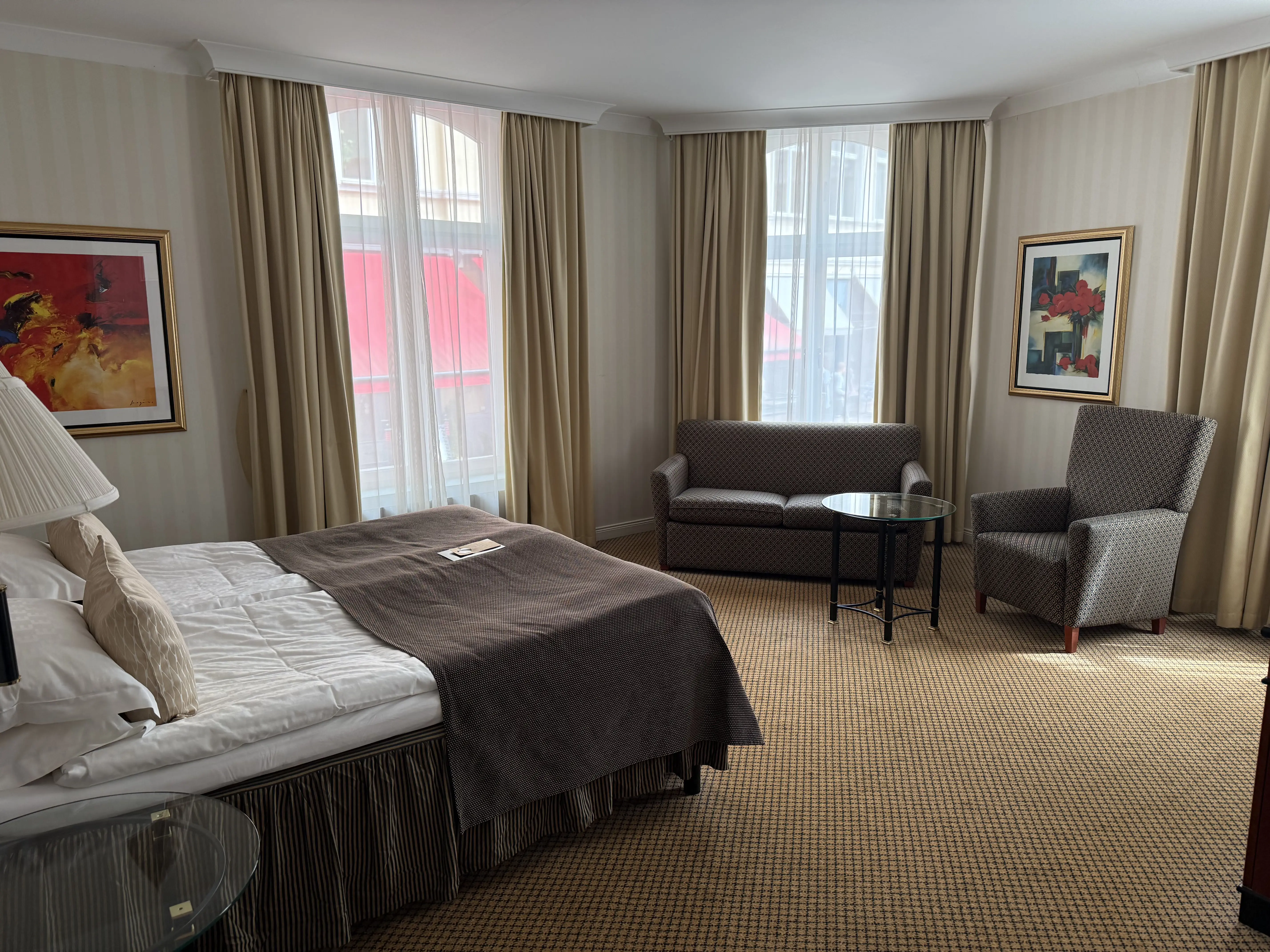 Modern hotel room with a cozy bed, stylish gray armchair, comfortable sofa, and round glass coffee table, accented by framed art and large windows with beige curtains.