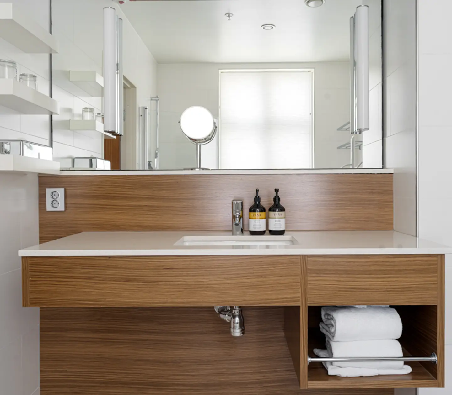 Modern bathroom vanity with wood finish, white countertop, and wall-mounted shelves, featuring a mirror, sink, toiletries, and neatly folded towels.