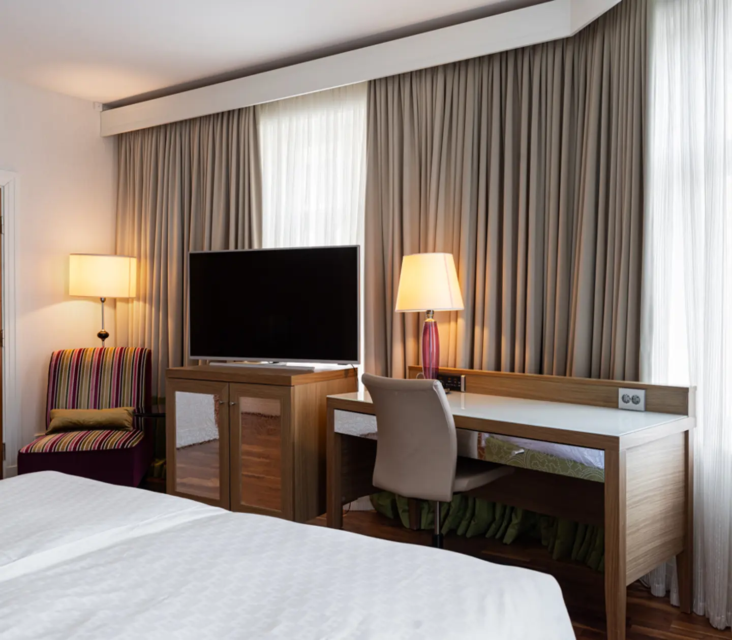 Modern hotel room with a cozy bed, wooden desk and chair, flat-screen TV on a stand, and elegant curtains. Two lamps provide warm lighting, complementing the striped armchair for a comfortable stay.