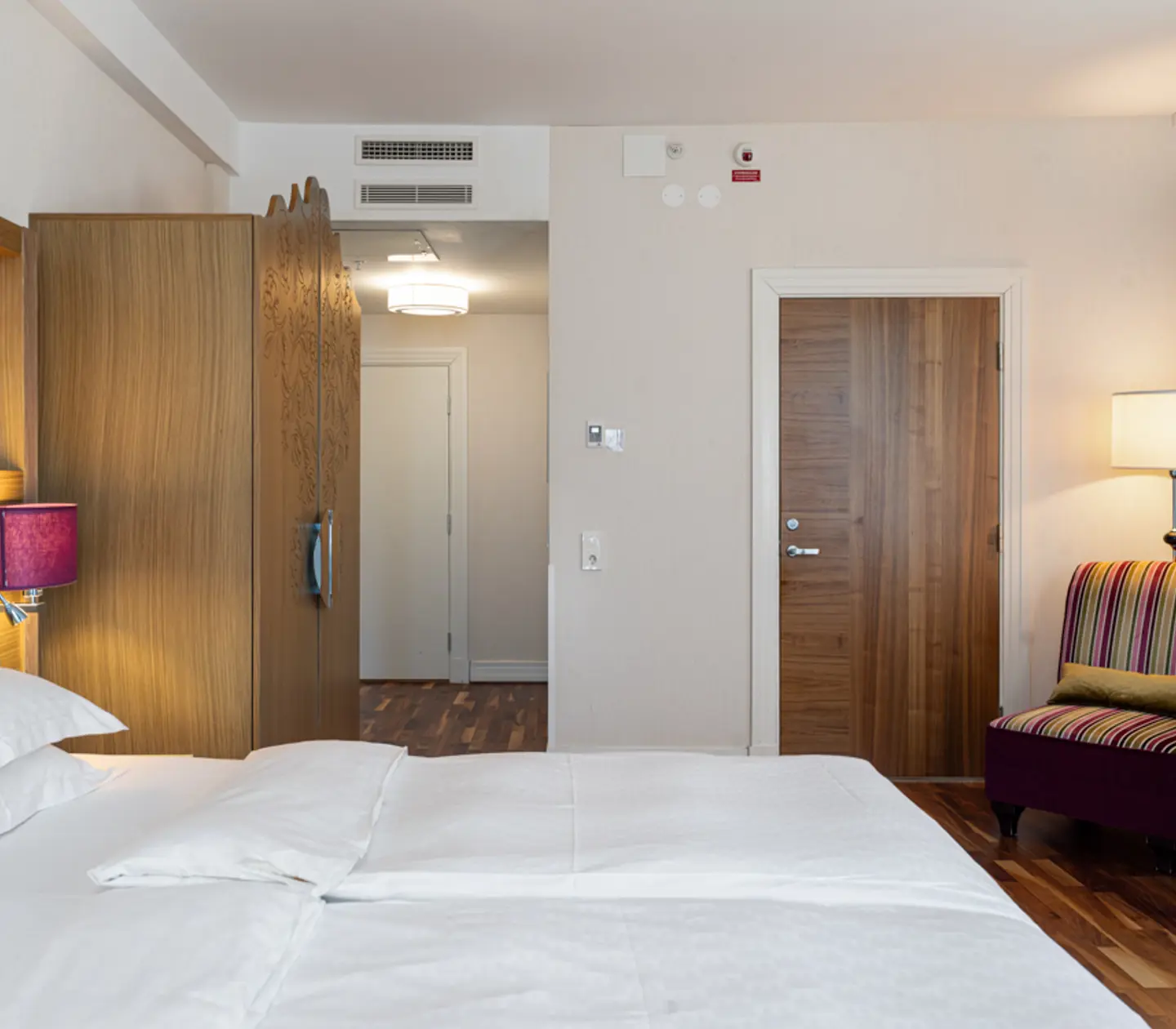 Hotel room interior with a cozy bed, wooden wardrobe, colorful striped chair, and ambient lighting from a table lamp.