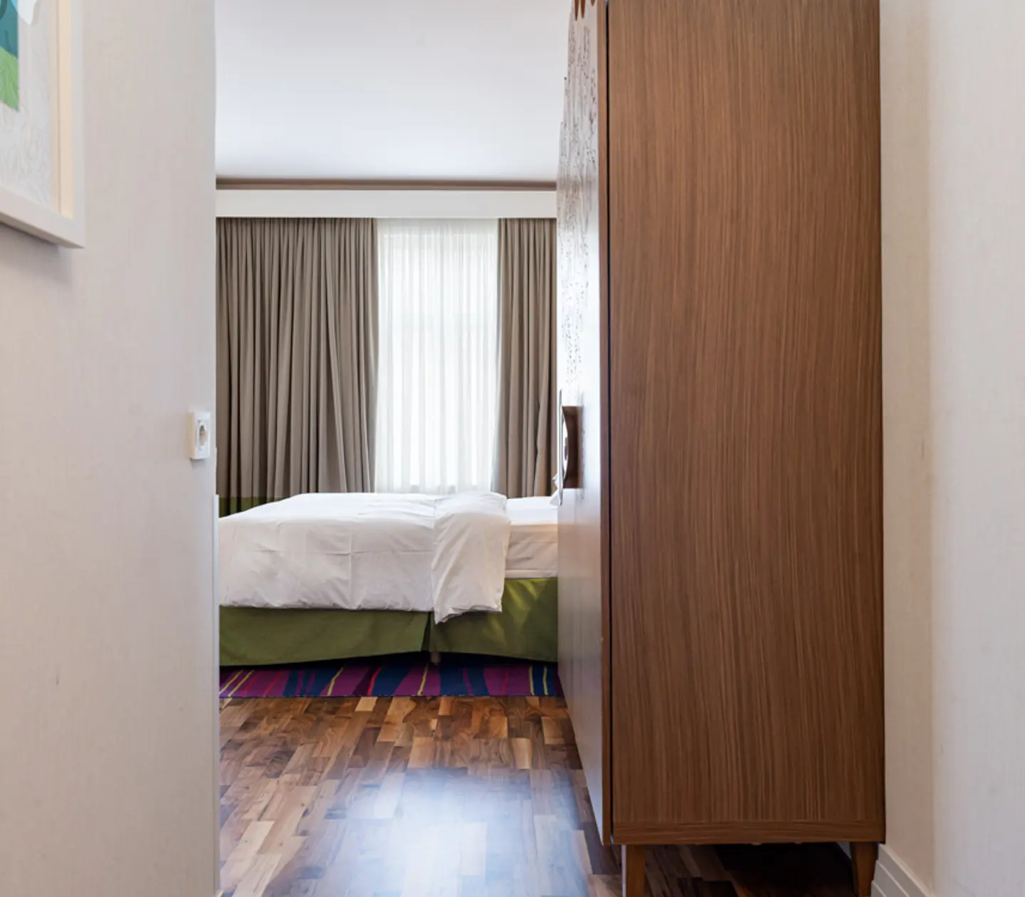 Modern hotel room entrance, showcasing a neatly made bed, elegant wooden wardrobe, large window with curtains, and polished hardwood floors.