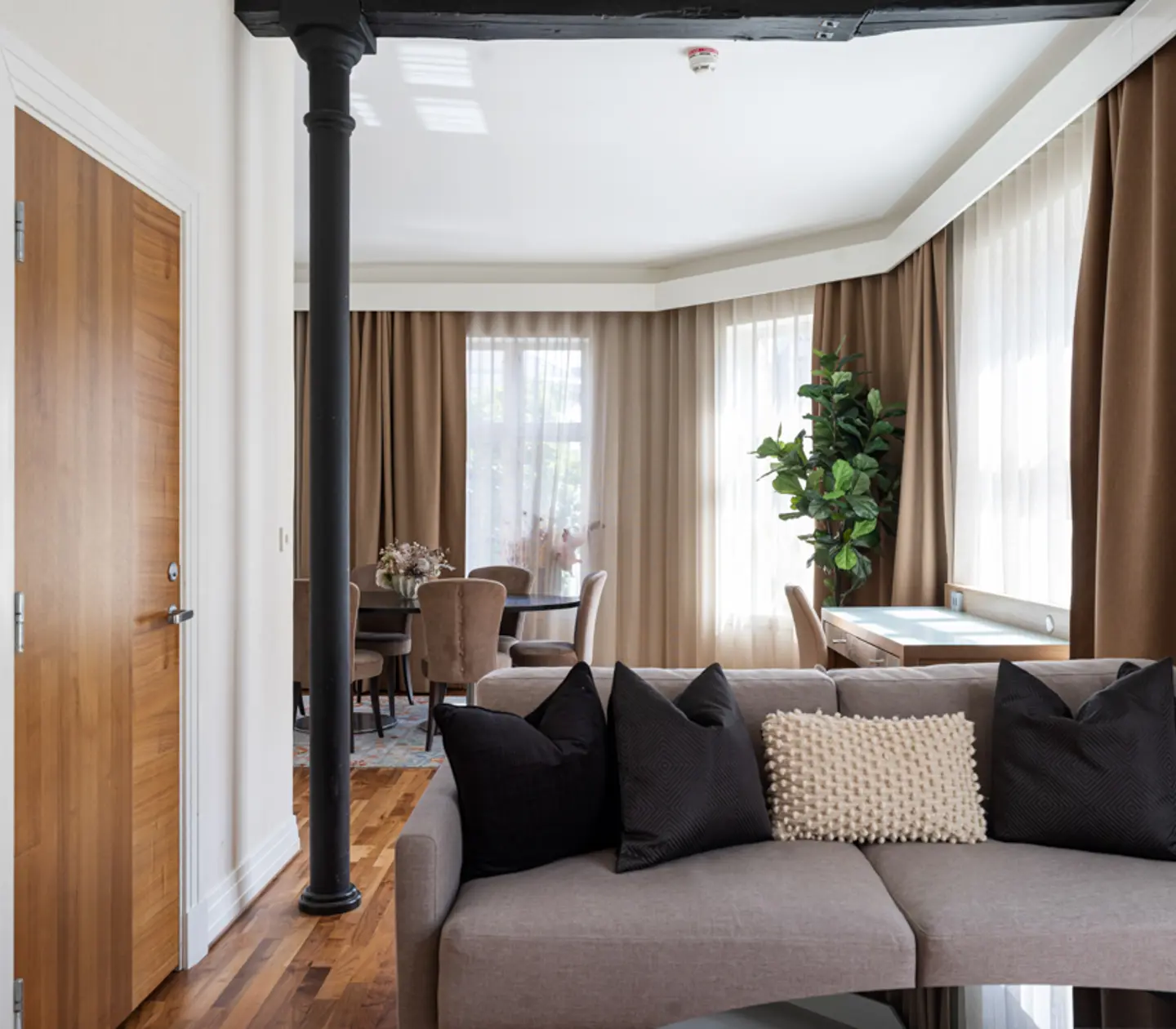 Stylish living room with a neutral-toned sofa, black and textured pillows, wooden door, exposed beam, dining table, chairs, and tall green plant, surrounded by large windows with brown curtains.