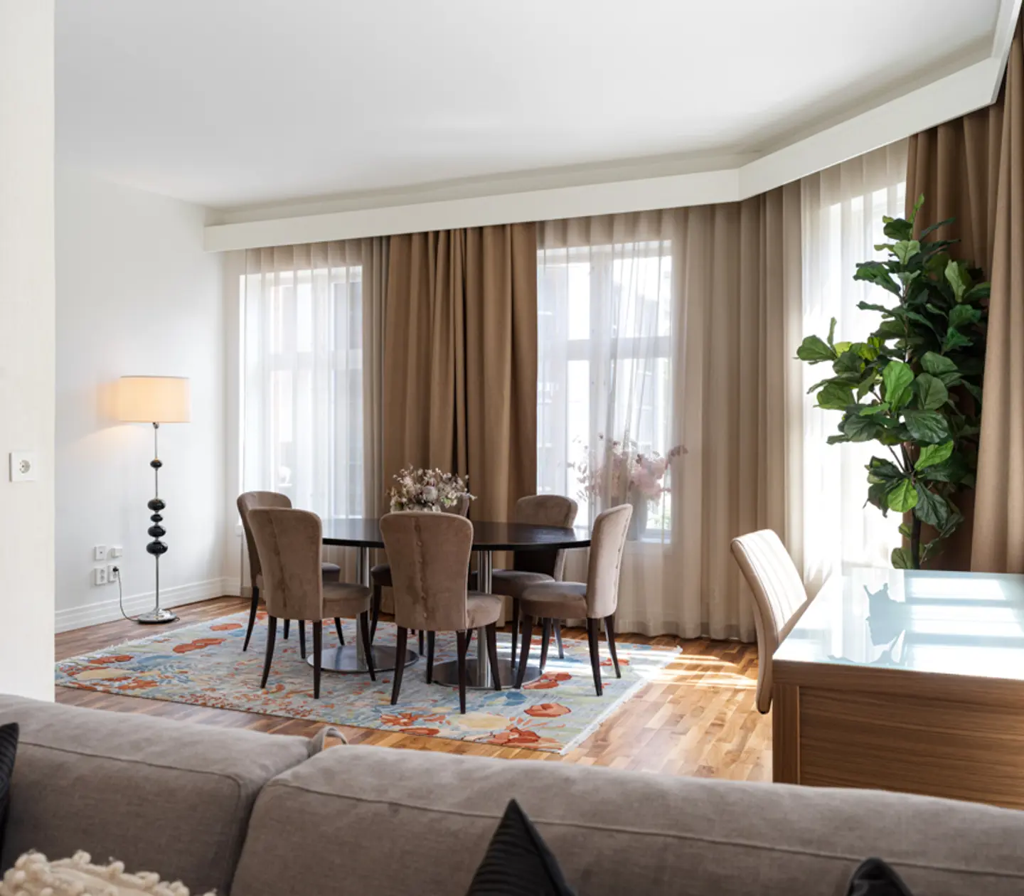 Elegant dining area with round table and chairs, large windows with beige curtains, floor lamp, indoor plant, wooden floors, and a decorative rug in a well-lit, modern living space.