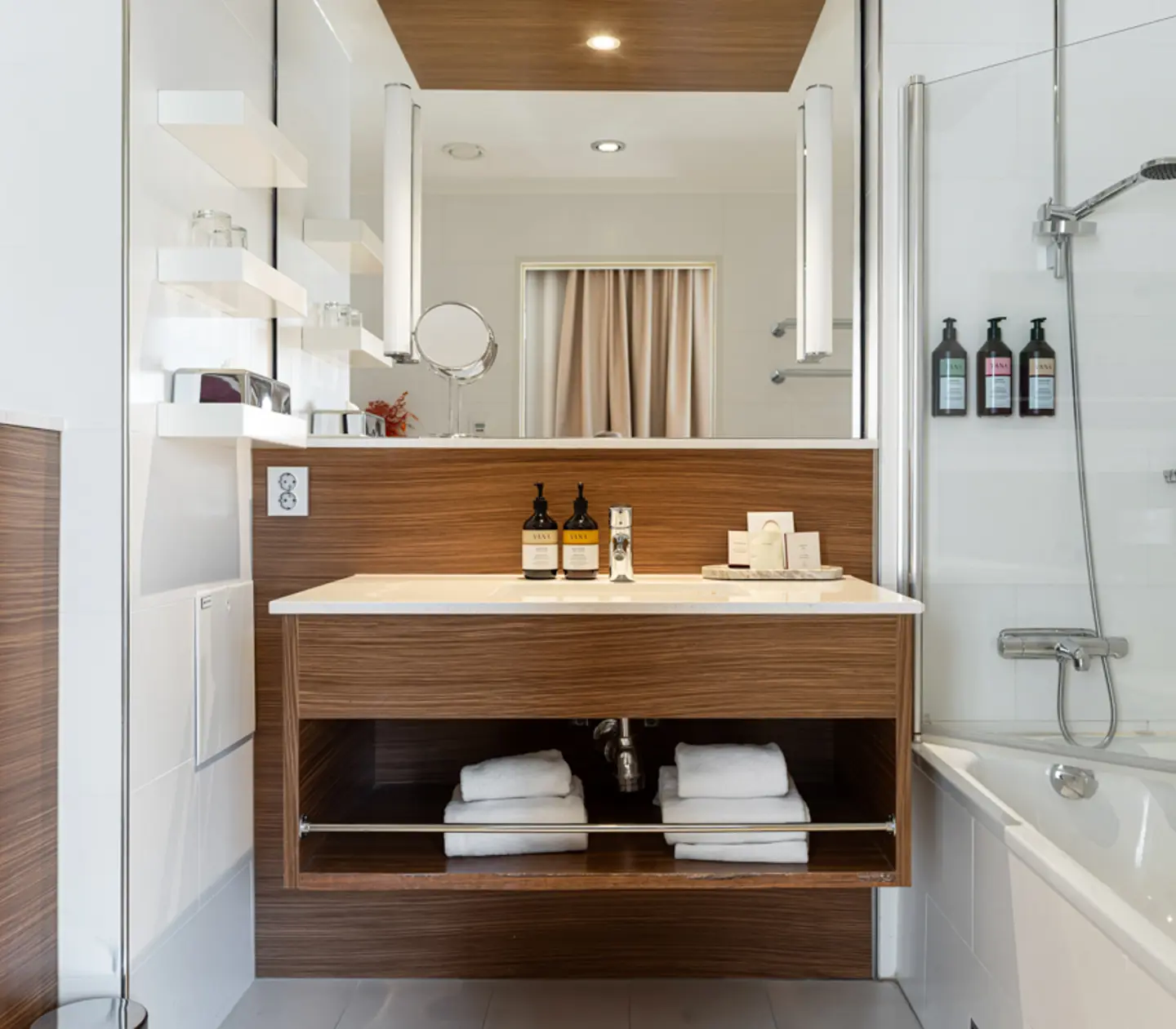 Modern bathroom with wooden vanity, under-sink towels, wall-mounted toiletries, and a white bathtub with a showerhead.