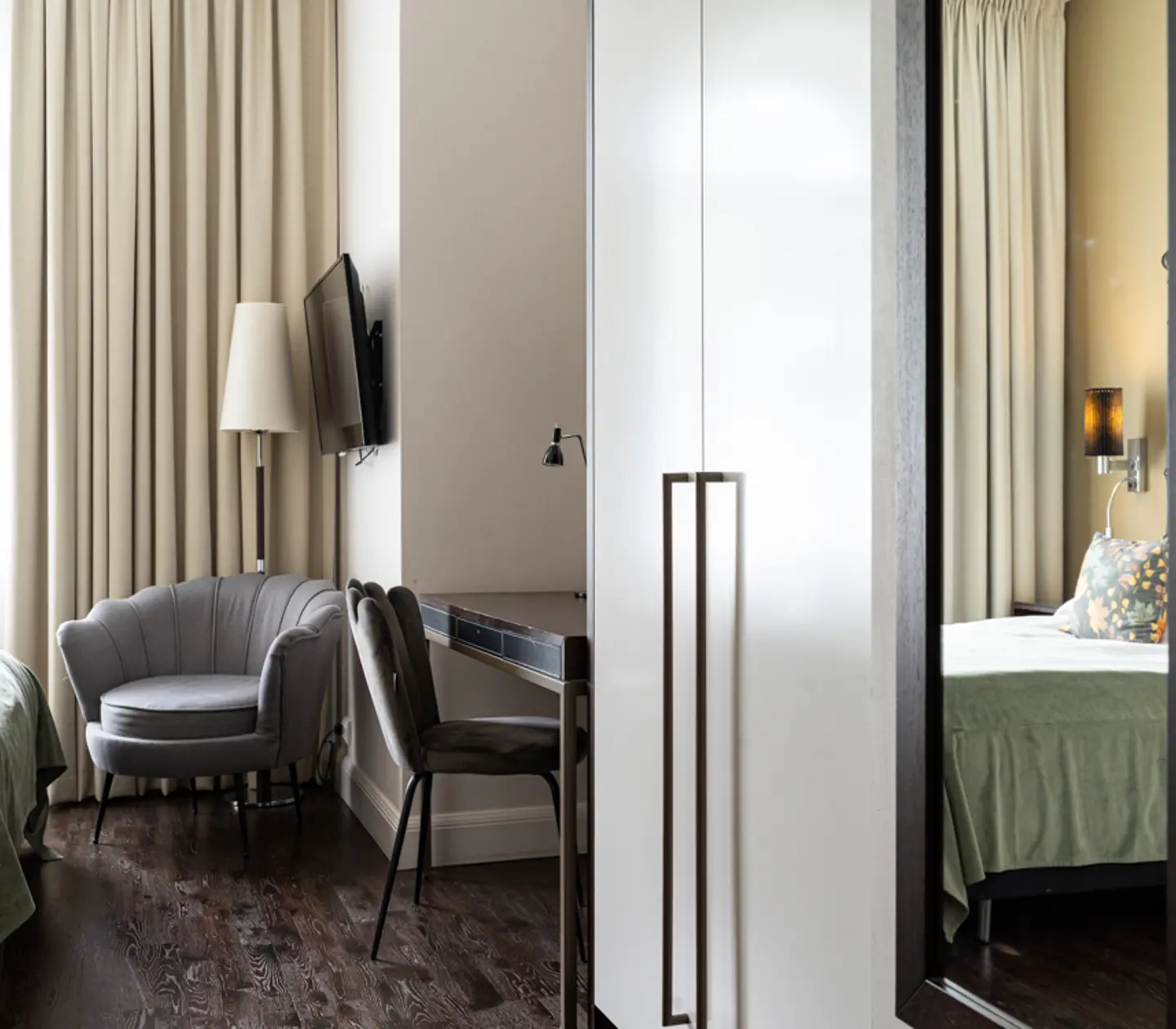 Modernt hotellrum med elegant inredning, inklusive bekväm gråfåtölj, skrivbord med stol, TV och spegel. Gardiner och en dubbelsäng med grön överkast syns, vilket skapar en avkopplande atmosfär.