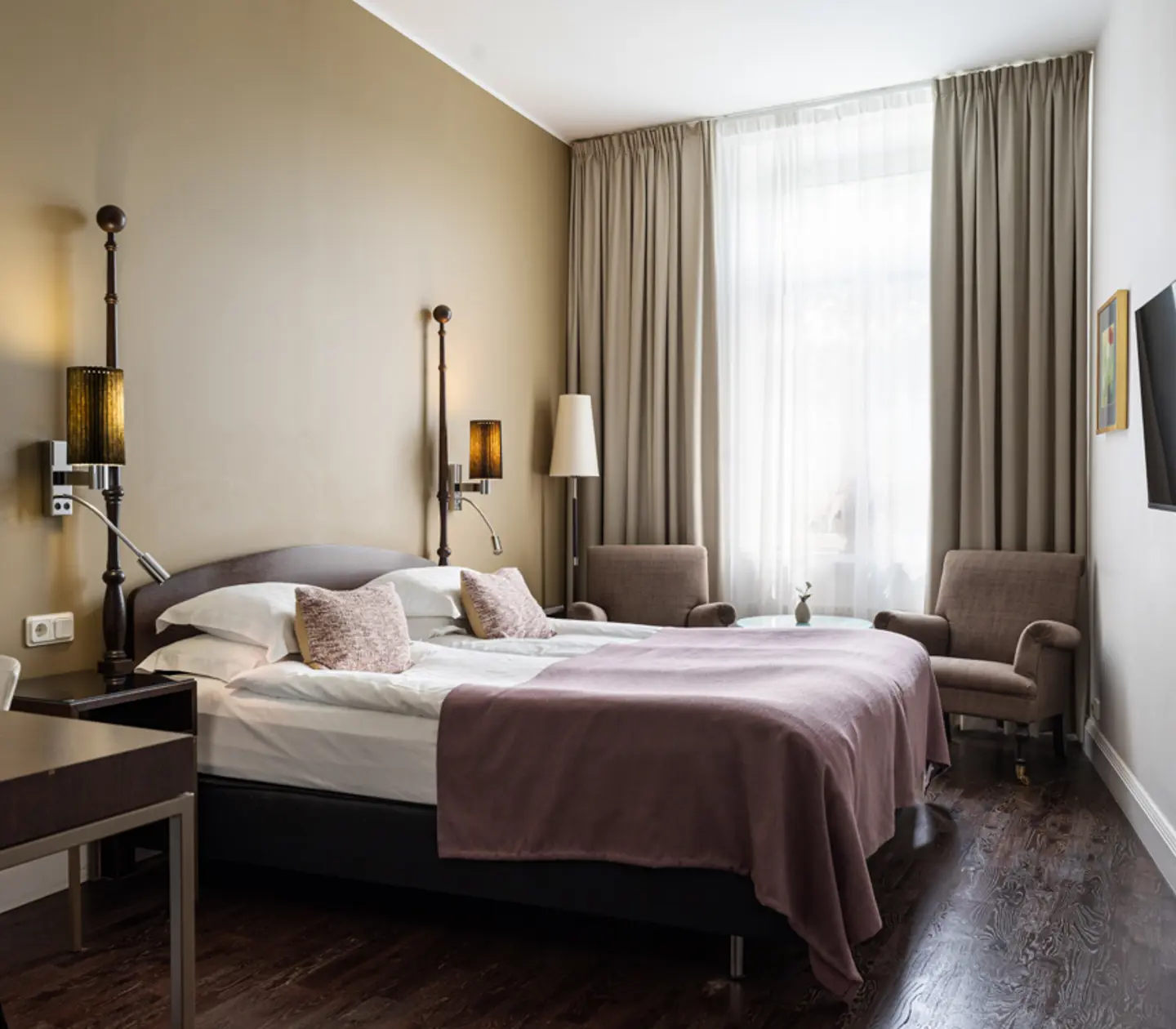 Ljust hotellrum med säng, lila fåtöljer och fönster