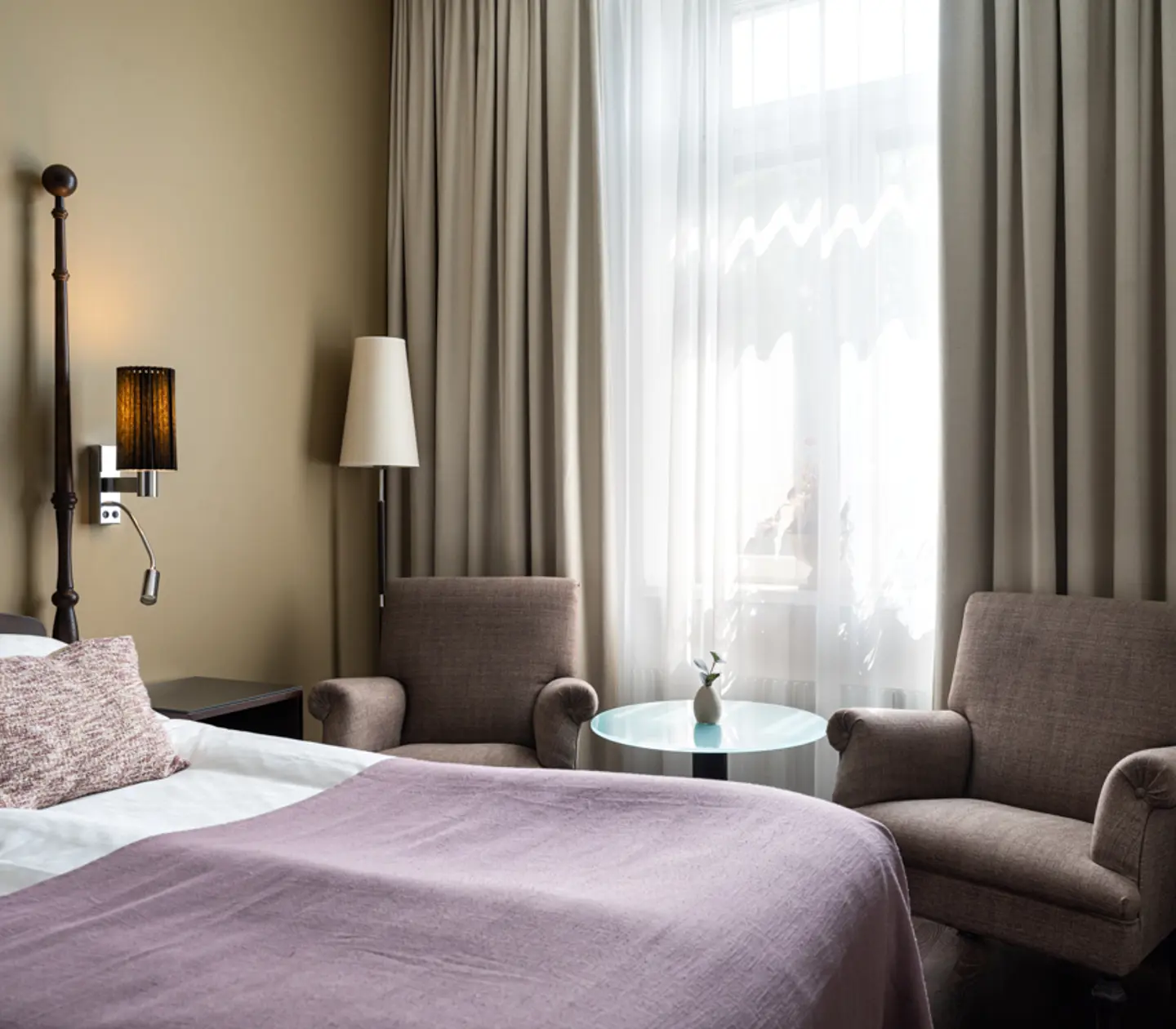 Ljust hotellrum med säng, lila fåtöljer och fönster