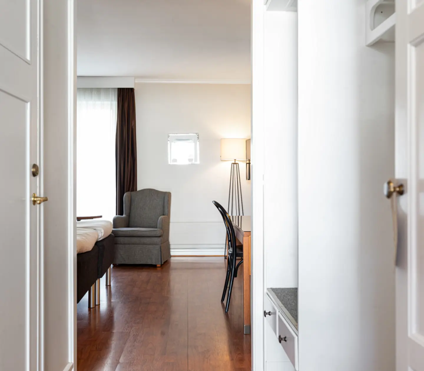 Modern hotellrum med öppna dörrar, bekväm fåtölj och skrivbord med stol, ljus inredning, trägolv.