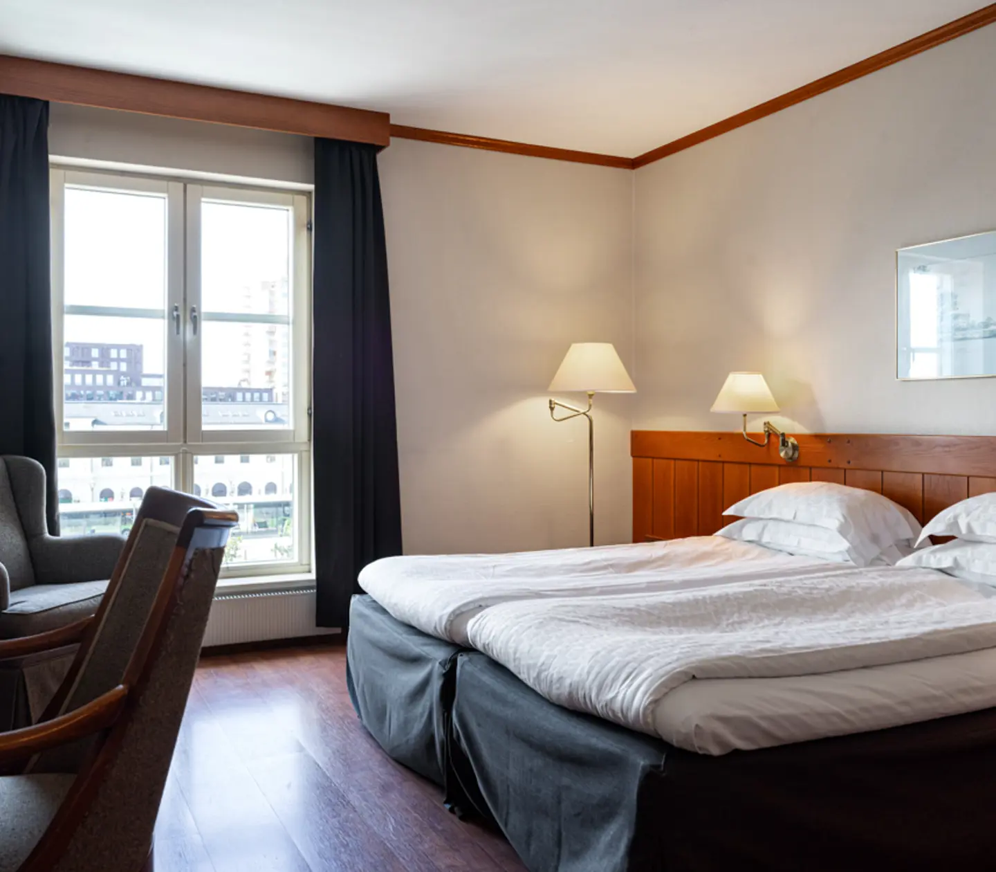 Hotellrum med dubbelsäng, fåtöljer, bord och stora fönster med utsikt, ljus inredning och stilrena möbler.