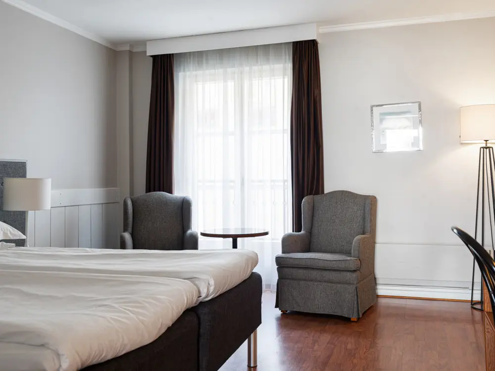 Ljust hotellrum med bekväm säng, två fåtöljer och stort fönster med gardiner, trägolv.
