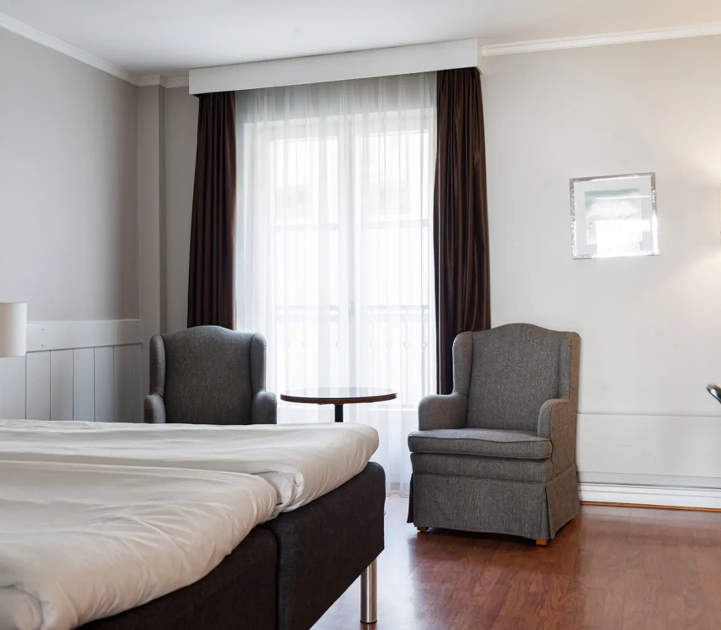 Ljust hotellrum med bekväm säng, två fåtöljer och stort fönster med gardiner, trägolv.
