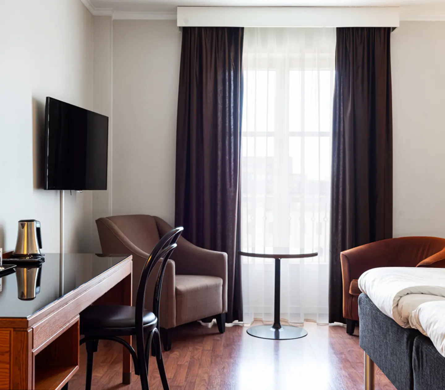 Modernt hotellrum med en bred säng, skrivbord, TV och två fåtöljer framför ett fönster med mörka gardiner.