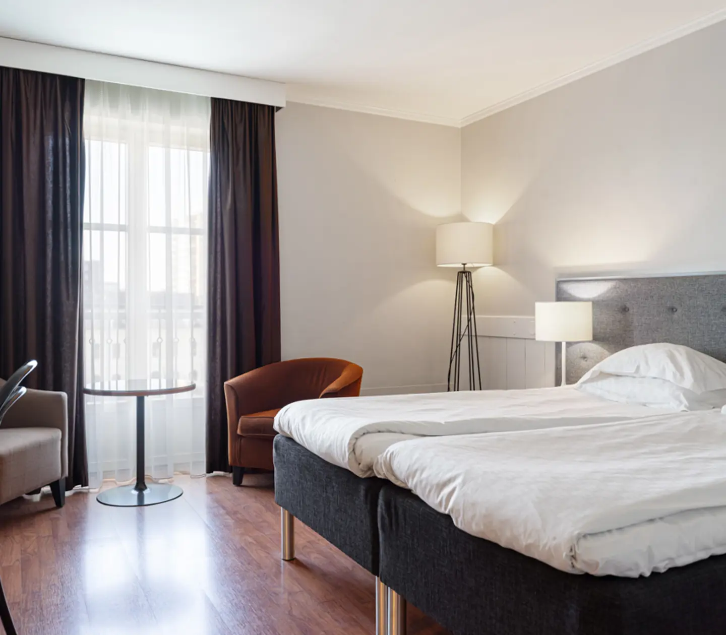 Modernt hotellrum med dubbelsäng, ljusa väggar, parkettgolv och stora fönster med mörka gardiner.
