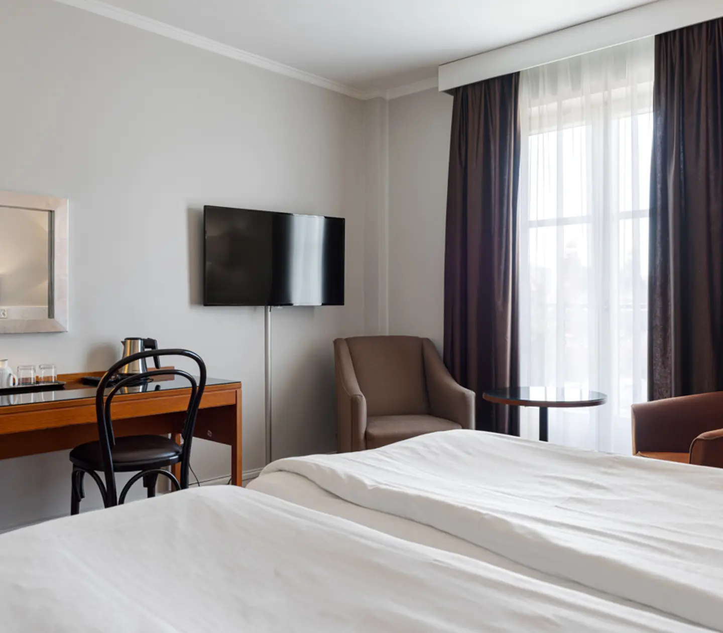 Hotellrum med en stor säng, skrivbord, stol och väggmonterad TV. Rummet har stora fönster med mörka gardiner och en fåtölj med ett litet bord.