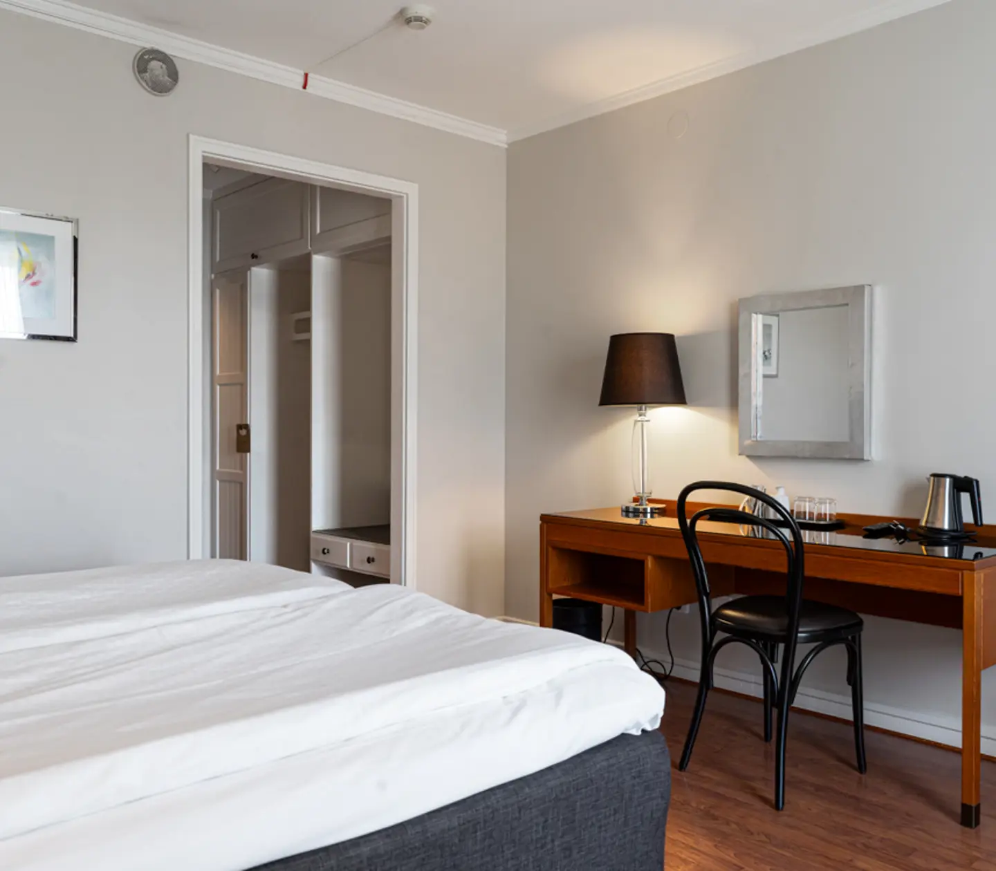 Ljust hotellrum med en bekväm säng, skrivbord, stol och bordslampa, perfekt för arbete eller avkoppling.