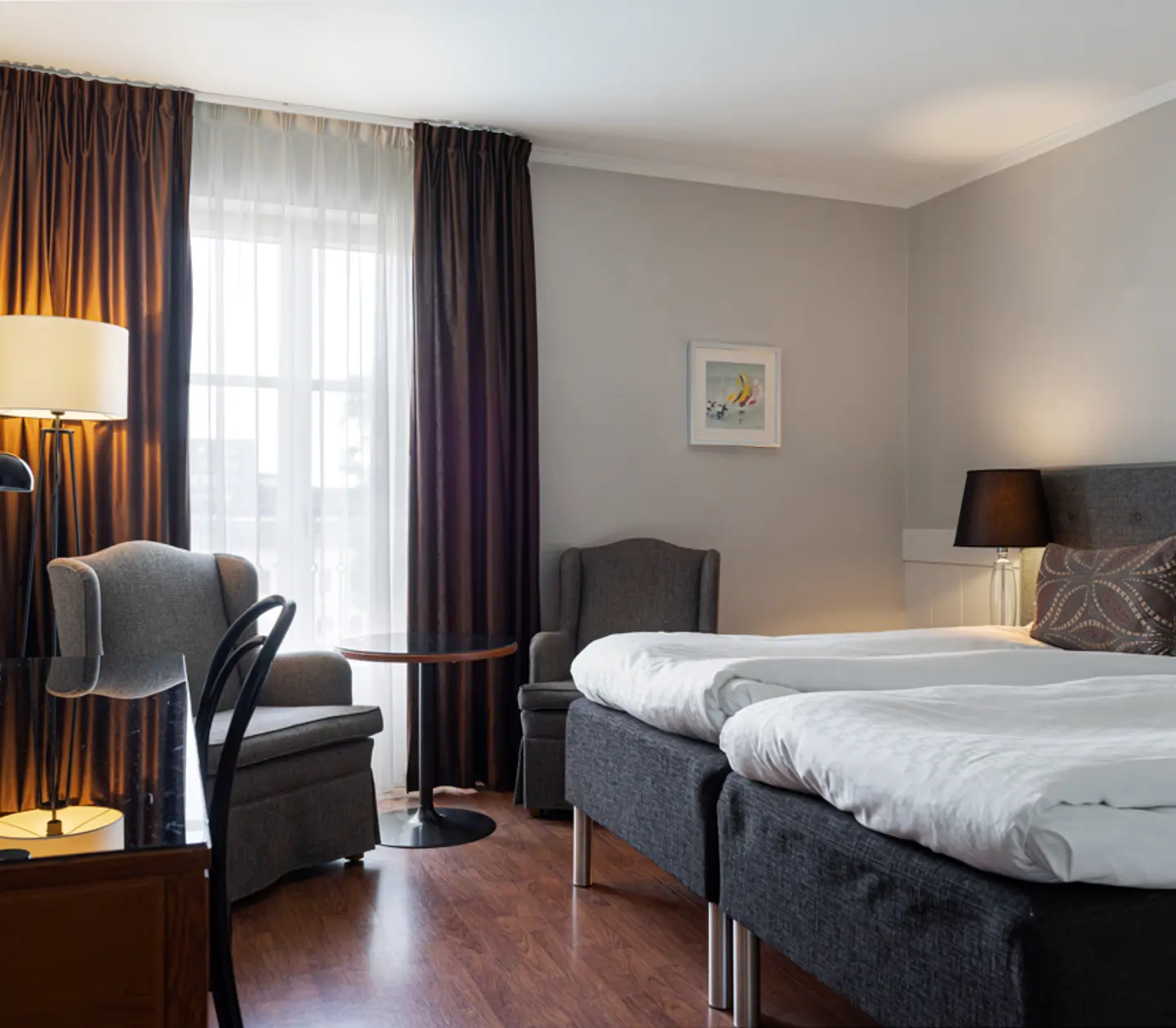 Ljust hotellrum med två enkelsängar, mörka gardiner och bekväma fåtöljer, beläget bredvid ett fönster för naturligt ljus.