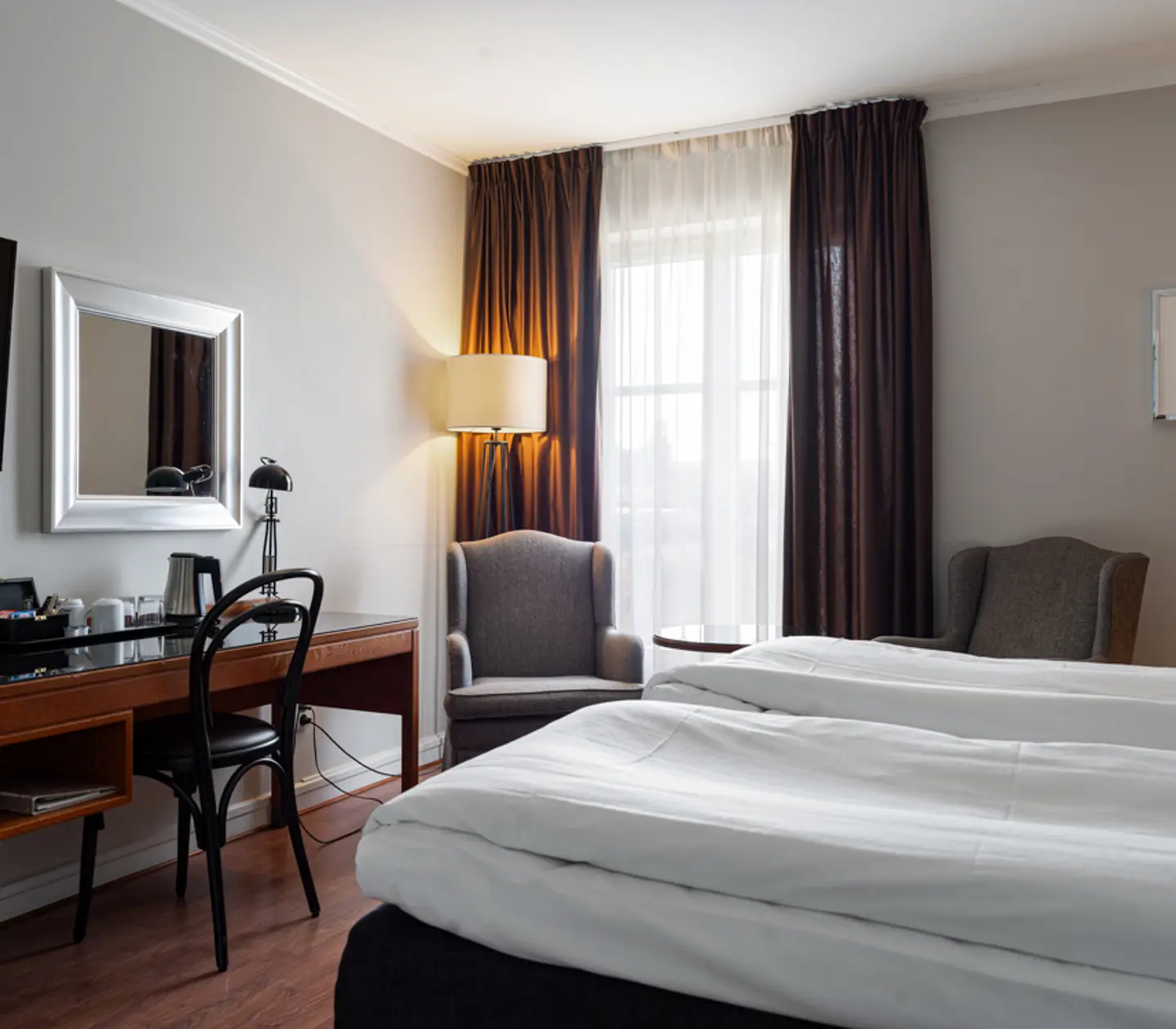 Ljust hotellrum med två sängar, skrivbord, TV och fåtöljer vid fönster med tjocka gardiner.