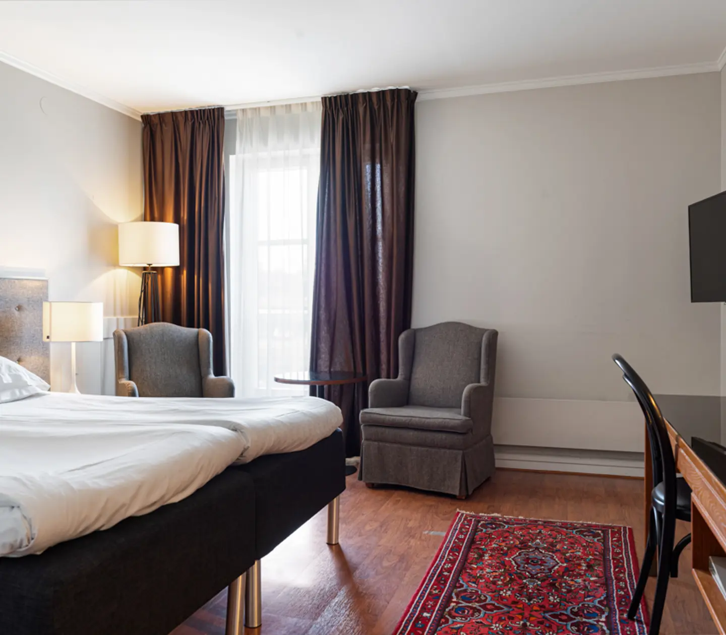 Modern hotellrum med dubbelsäng, grå fåtöljer, skrivbord och TV. Stora fönster med mörka gardiner och en röd matta på trägolv.