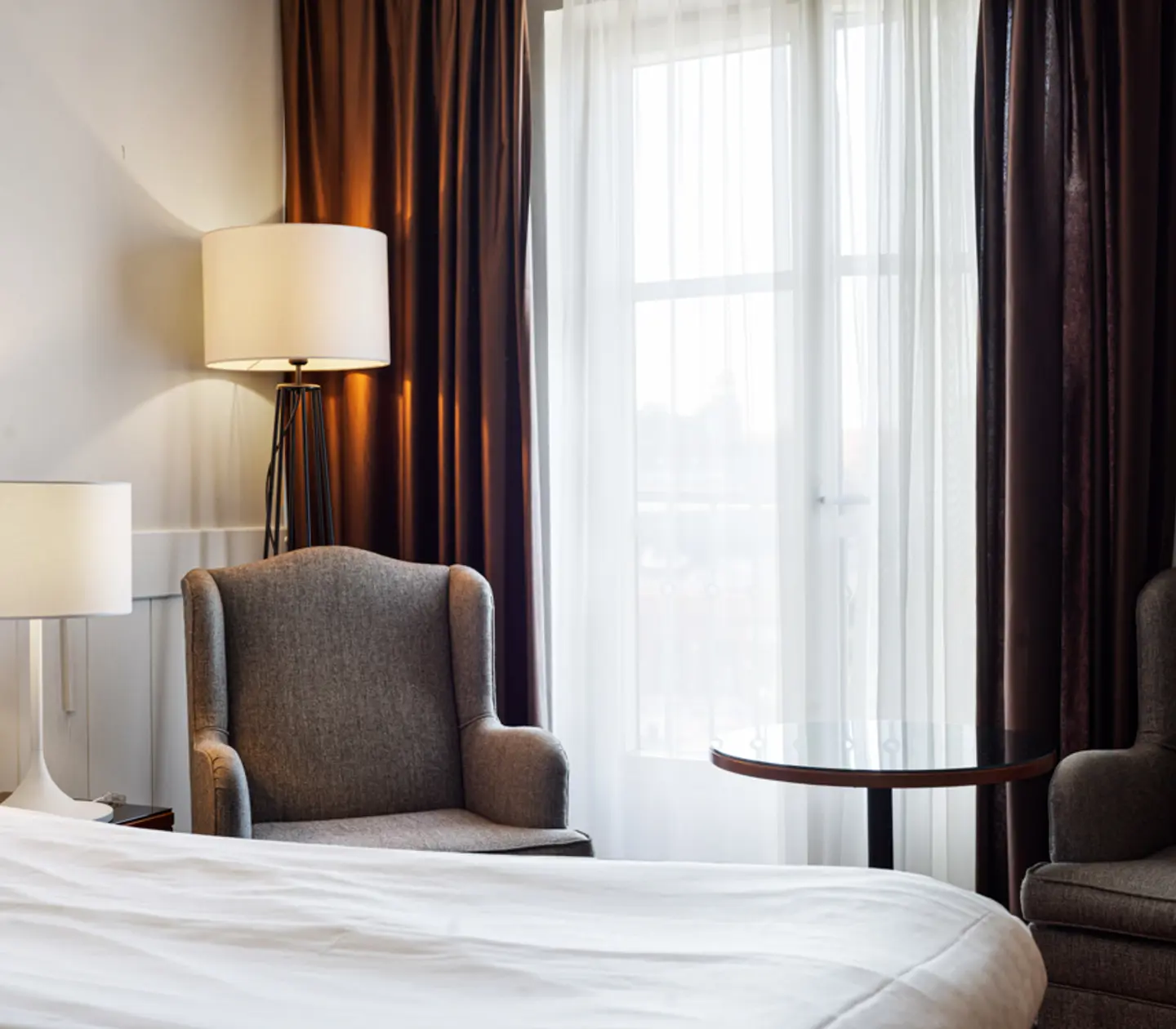 Modernt hotellrum med två grå fåtöljer, vita sängkläder och två bordslampor, delvis skymt av mörka och vita gardiner vid ett stort fönster.