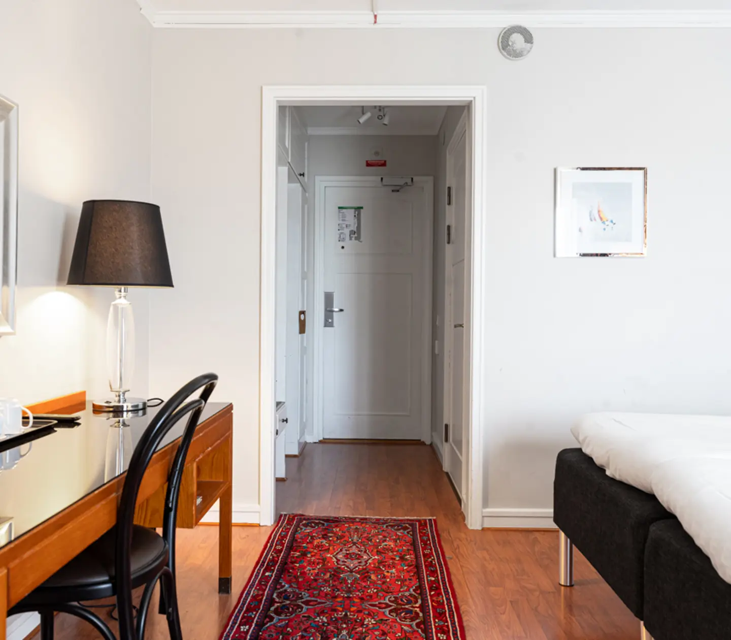 Hotellrum med enkel inredning, skrivbord med svart stol och lampa, säng med vit sängkläder och röd orientmatta på trägolv.