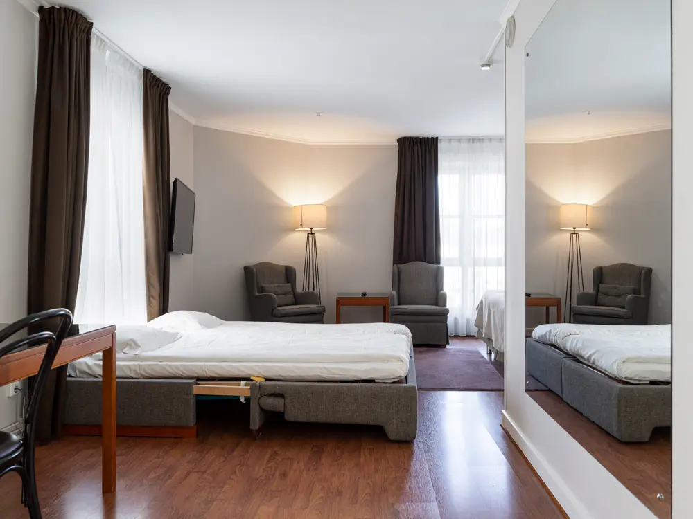 Modernt hotellrum med grå färgsättning, dubbelsäng, fåtölj, spegel och TV, samt stort fönster med mörka gardiner.