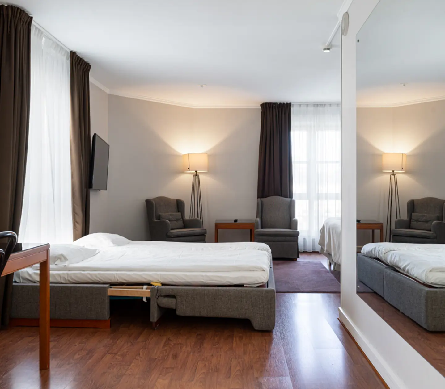 Modernt hotellrum med grå färgsättning, dubbelsäng, fåtölj, spegel och TV, samt stort fönster med mörka gardiner.