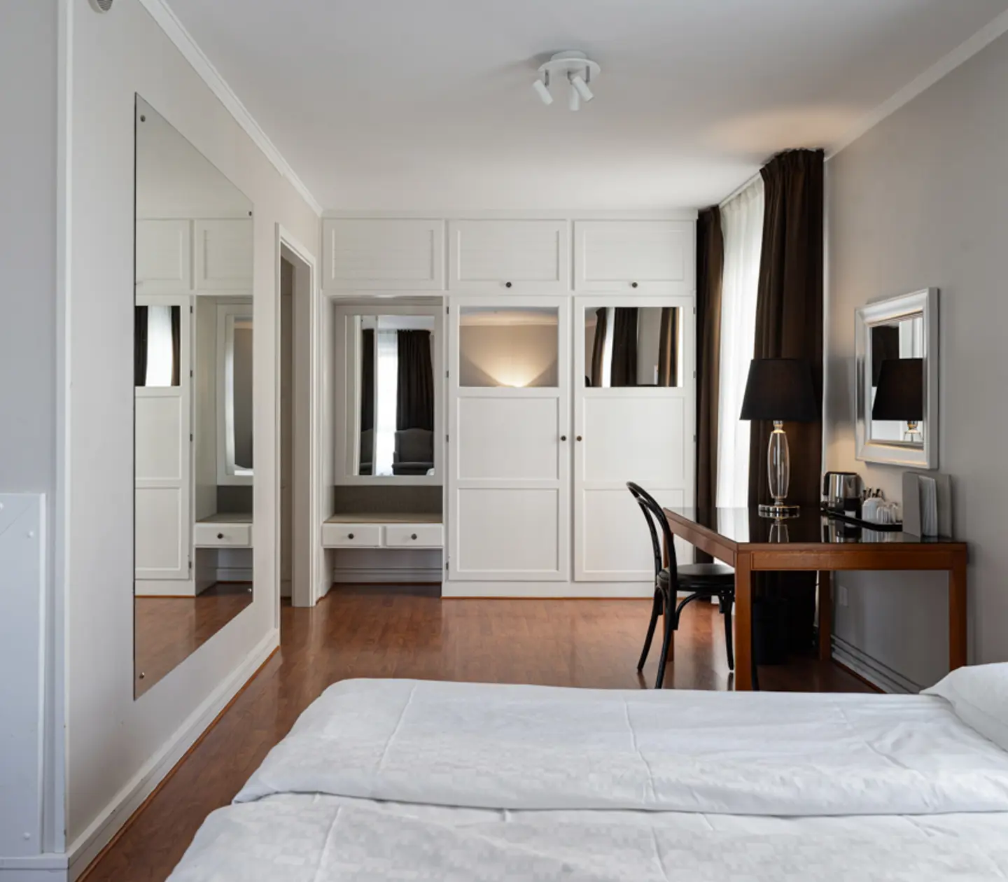 Modernt hotellrum med vit säng, väggspegel, skrivbord och garderober.