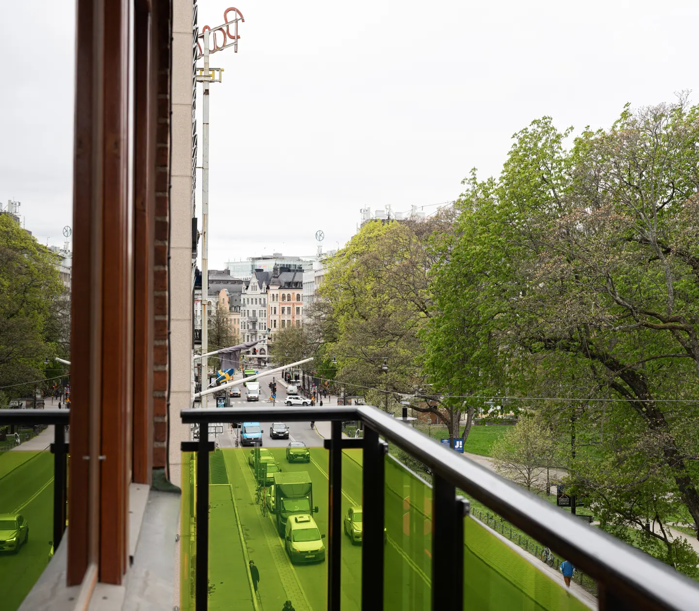 Utsikt från balkong över en livlig stadsgata kantad av grönskande träd i centrala Stockholm, med historiska byggnader i bakgrunden.