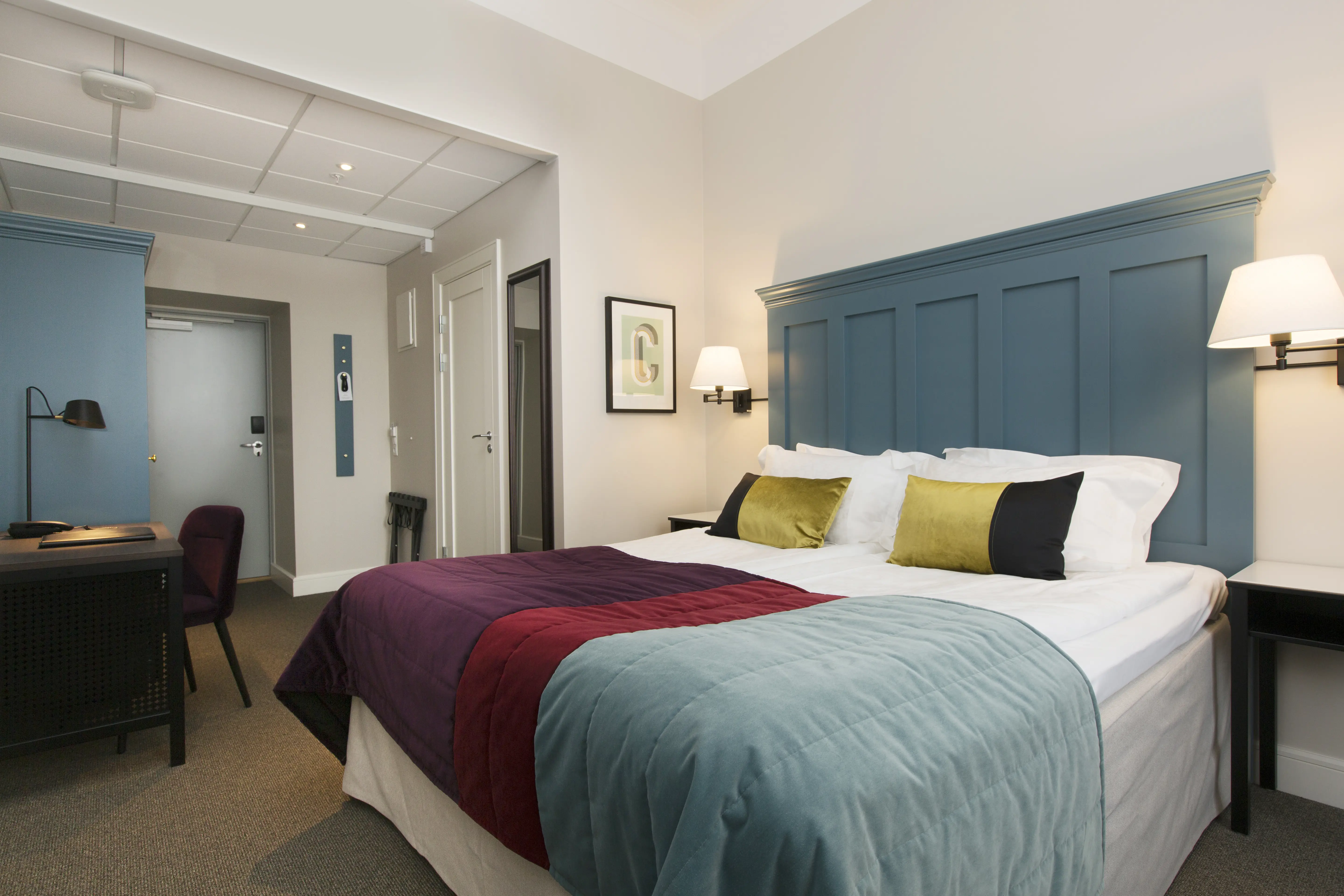 Bright hotel room with bed, large blue headboard and desk