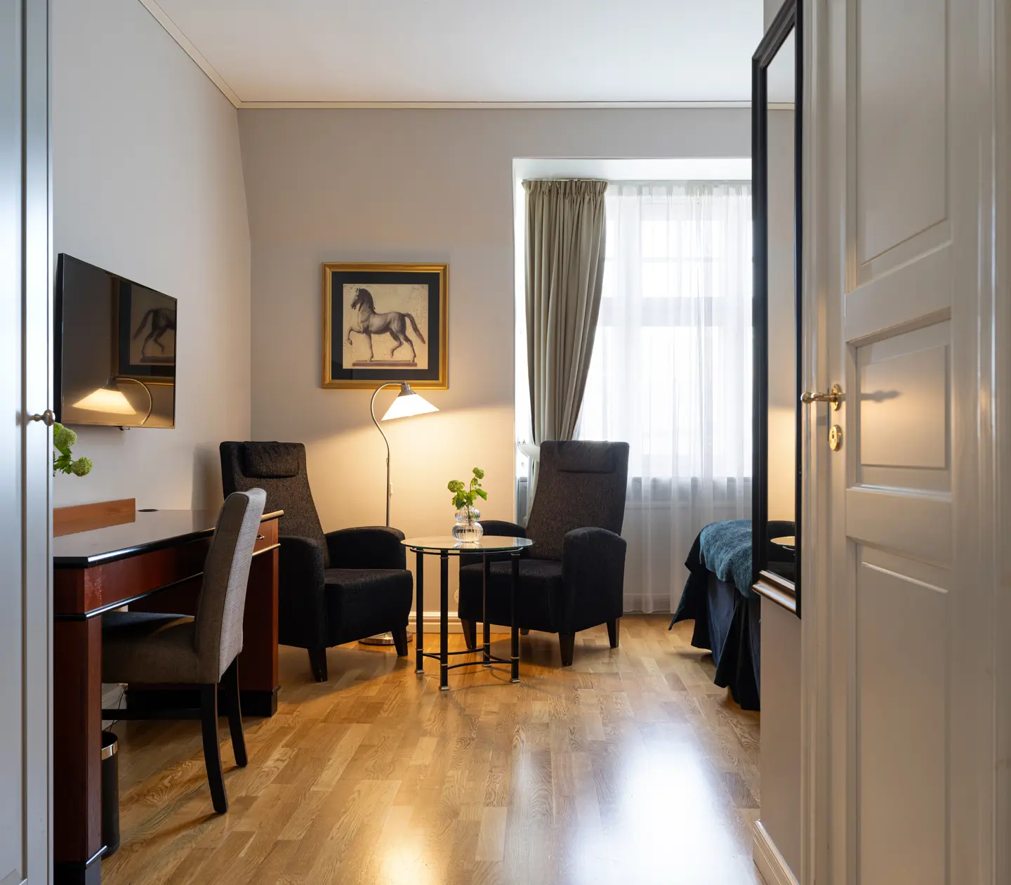 Elegant hotellrum med trägolv, två fåtöljer, skrivbord och platt-TV. Ljus fönsterbelysning med gardiner ger en sofistikerad känsla.