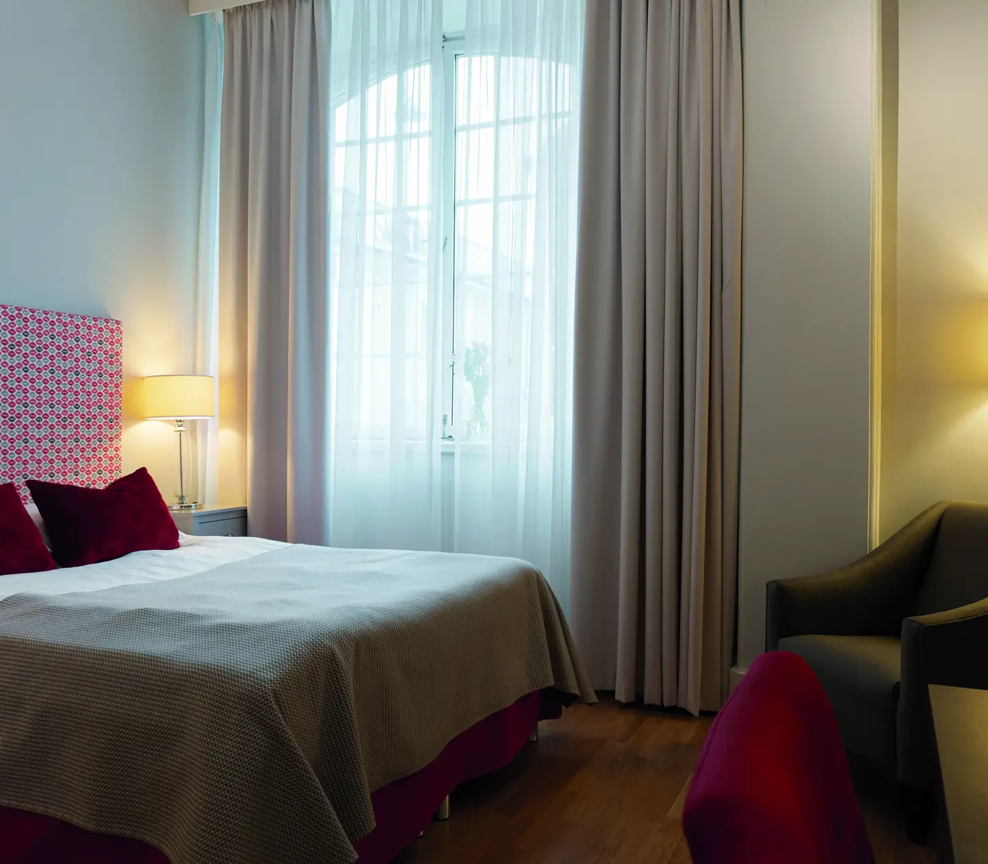 Modernt hotellrum med dubbelsäng, färgglad sänggavel och vita gardiner. Varm belysning, fåtölj och skrivbord i hörnet.