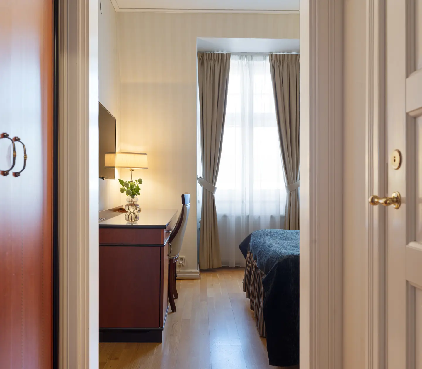 Hotellrum med mjuk belysning, träpaneler och en säng bredvid fönster med ljusa gardiner. En skänk med lampa och växt pryder rummet.