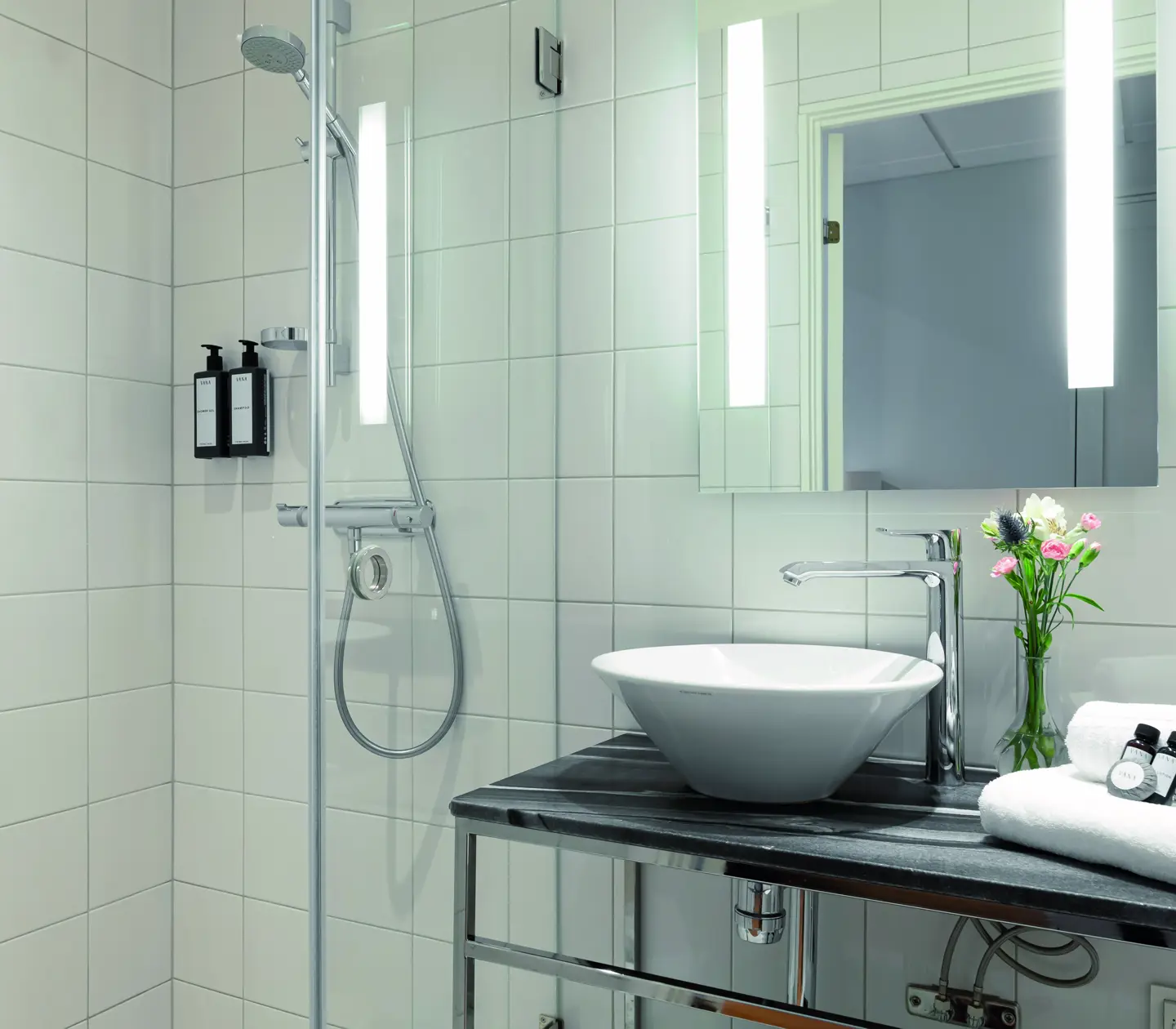 Modernt badrum med duschkabin, spegel med belysning, handfatsskål och blommor på en bänk tillsammans med vikta handdukar och hygienprodukter.