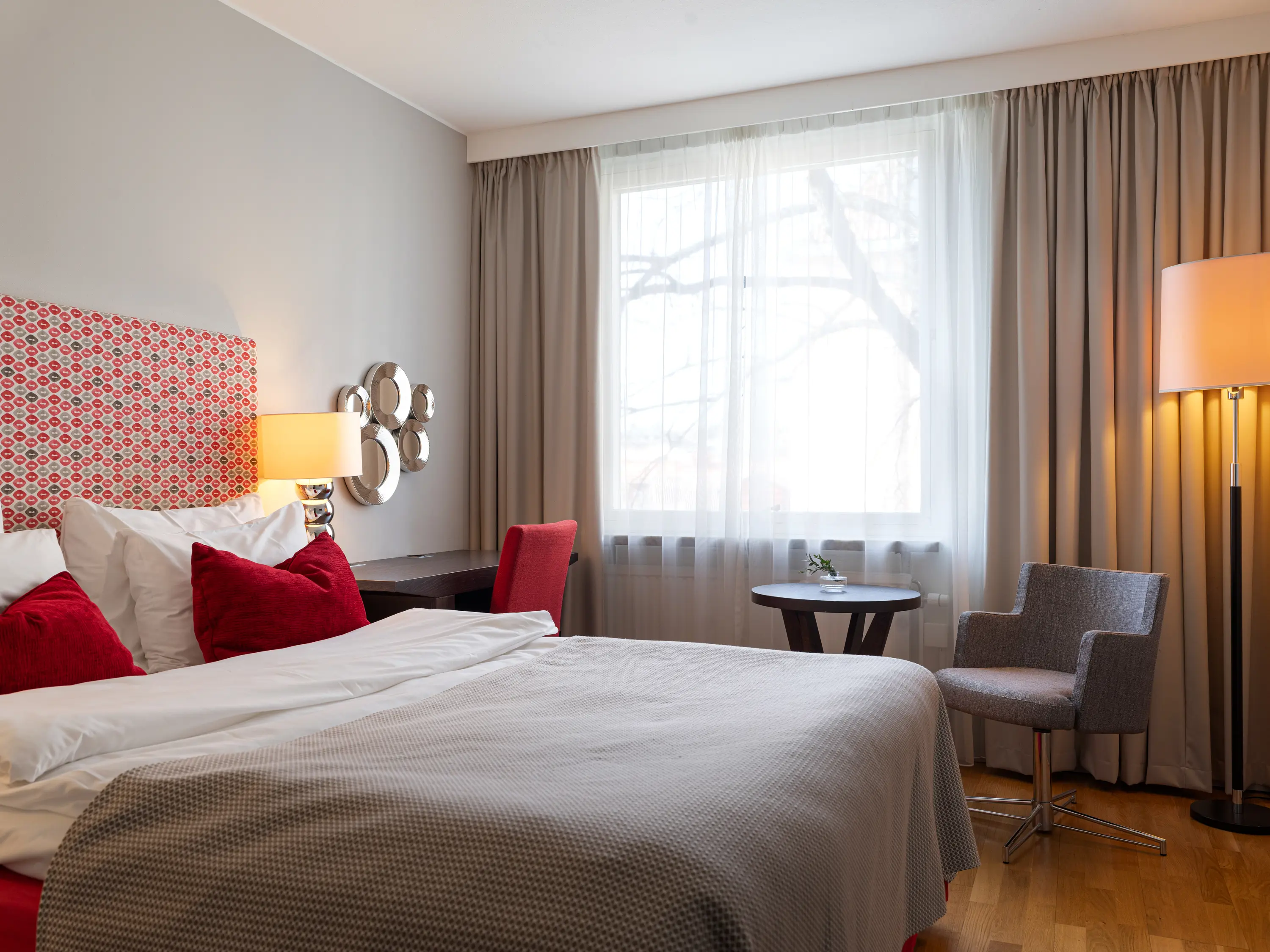 Modernt hotellrum med dubbelsäng, röda kuddar och elegant inredning, inklusive skrivbord, fåtölj och golvlampa, bredvid ett stort fönster med gardiner.