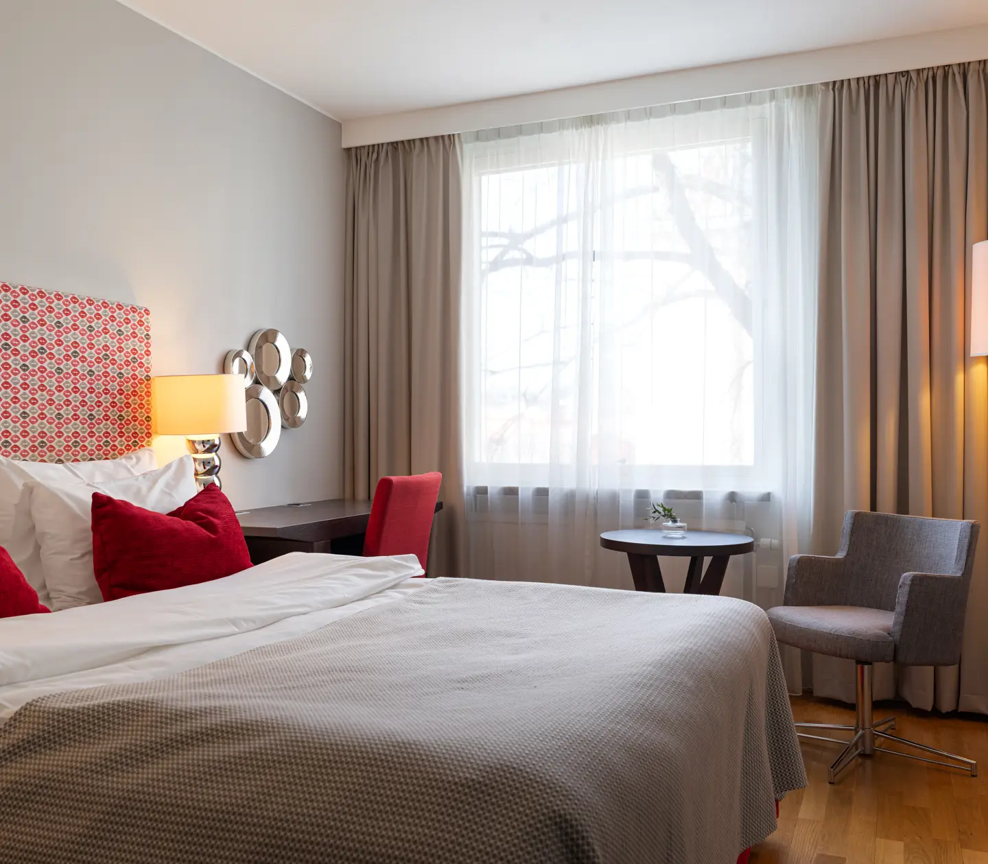 Modernt hotellrum med dubbelsäng, röda kuddar och elegant inredning, inklusive skrivbord, fåtölj och golvlampa, bredvid ett stort fönster med gardiner.
