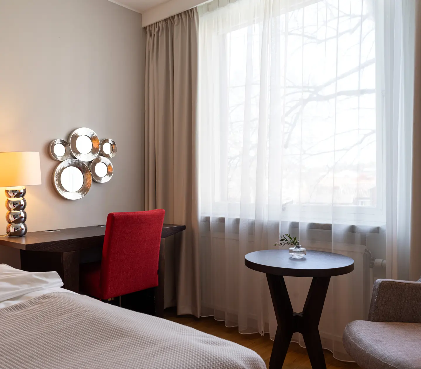 Modernt hotellrum med röd stol, skrivbord och säng nära fönster med vita gardiner.