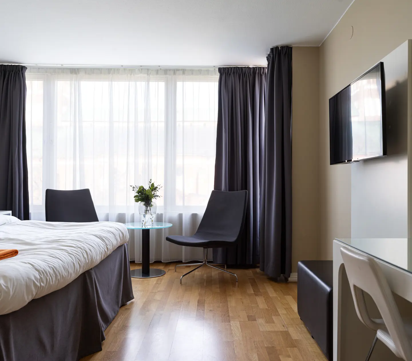 Modernt hotellrum med dubbelsäng, parkettgolv, två svarta fåtöljer och en väggmonterad TV.