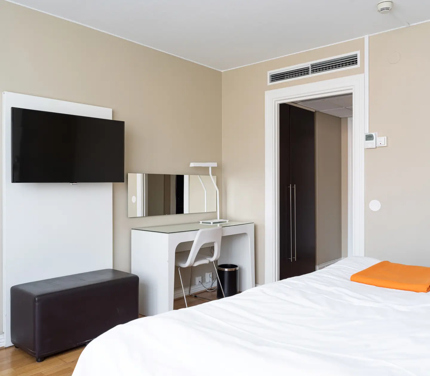 Modernt hotellrum med plattskärms-TV, skrivbord, spegel och en säng med vit och orange sängkläder i minimalistisk inredning.