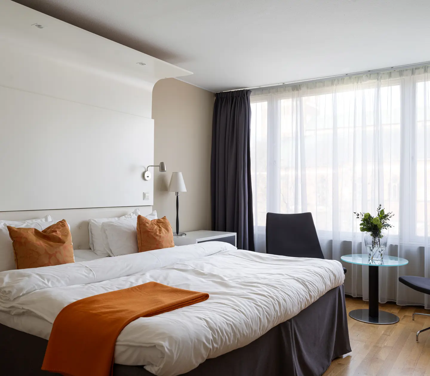 Hotellrum med en stor bäddad säng, vita lakan och orange kuddar, bredvid ett fönster med gardiner. Det finns ett litet bord med en blomvas och två mörka stolar.